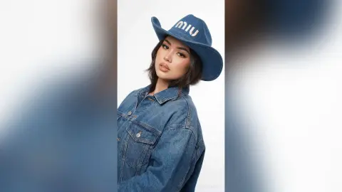 Stylish woman in a blue denim jacket and Miu Miu cowboy hat posing against a white background.