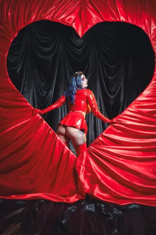KimLennoxc poses in a red latex outfit and blue hair, peeking through a large red heart-shaped curtain opening against black drapes.