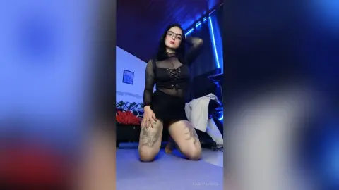 Young woman with glasses in a black sheer lace top and mini skirt posing in a moody blue lit bedroom.