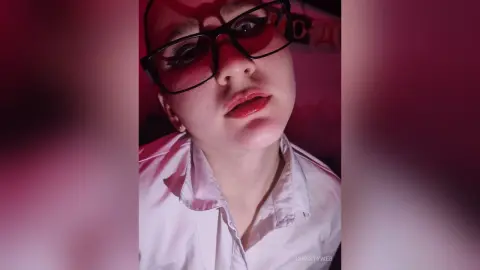 ChristyWeb wearing black glasses and a white button-down shirt, looking directly at the camera with red lipstick.