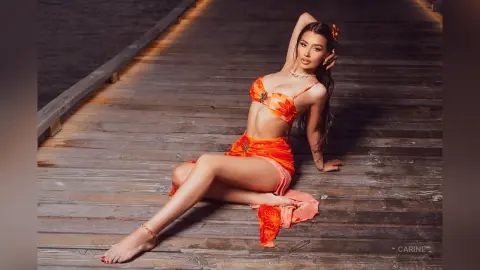 Carine sits on a wooden pier wearing an orange lace bikini set and a matching wrap, posing with one hand in her long dark hair.