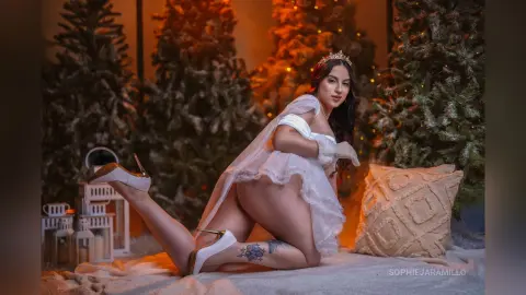 Glamorous woman in a white lace outfit and tiara posing in a cozy, festive winter setting with Christmas trees.