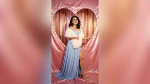 Smiling woman in a light blue evening gown and white fur stole posing in a pink heart-shaped photo booth.