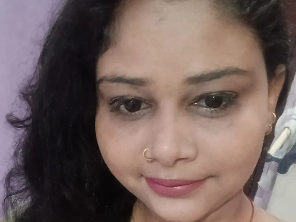 CurvySoniya smiles close up, showing her dark wavy hair, nose ring, and pink lipstick against a plain purple background.