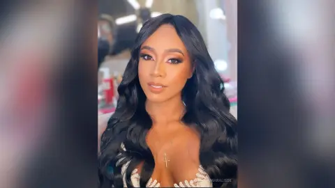 Glamorous woman with long wavy black hair and glowing makeup wearing a white lace top in a soft-lit setting.