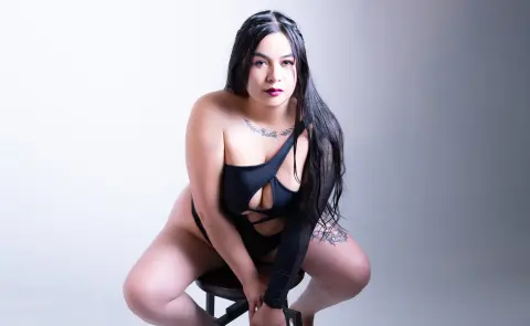 HanahMiler poses in a black cut-out bodysuit, squatting on a stool against a plain white background with her long black hair down.