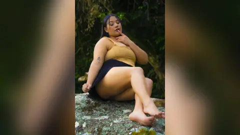 Curvy woman in a tan bodysuit sitting on a mossy rock in a lush green forest setting.