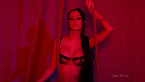 Glamorous woman in black lingerie posing between ropes under moody red studio lighting.