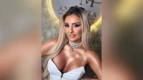 Anita poses with long blonde hair and a silver choker, wearing a white zip-up top that shows deep cleavage.