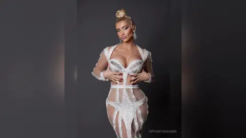 TiffanyAddams poses in a white, sheer, beaded bodysuit that shows off her cleavage and curves against a dark grey background.