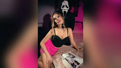 BeaNix reclines on a bed wearing a black lace bra and pink skirt, posing next to a person in a Scream mask.
