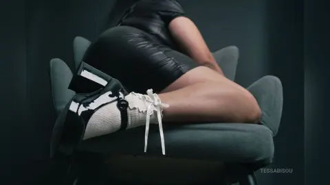 Woman in a black leather mini dress and white ruffled socks sitting on a grey chair in a moody setting.