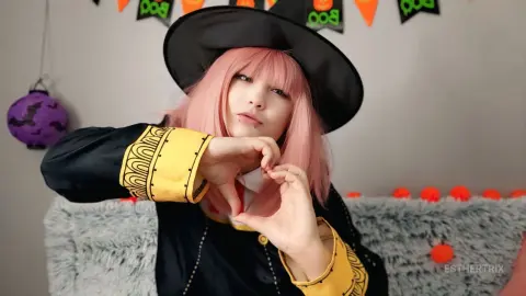 EstherTrix wears a black witch costume and hat while making a heart shape with her hands against a Halloween background.