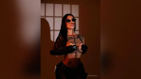 OrianaWill poses in sunglasses and black gloves, wearing a sheer black top that shows her chest tattoos and nipples.