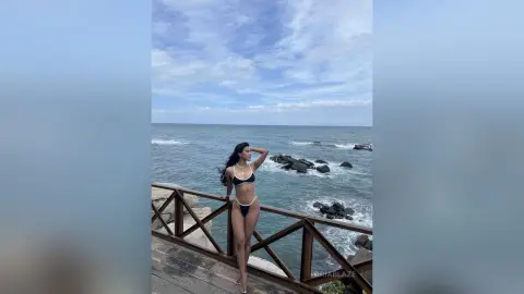 Slim woman in a black bikini posing on a wooden pier overlooking a vast blue ocean under a cloudy sky.