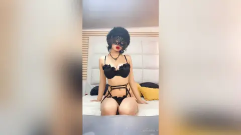 Woman in black lace lingerie sitting on a bed in a bright bedroom setting.