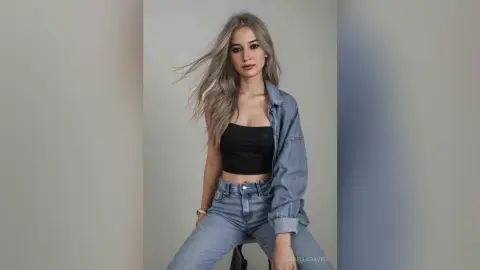 Young woman with long blonde hair wearing a black crop top and denim jacket in a minimalist studio setting.