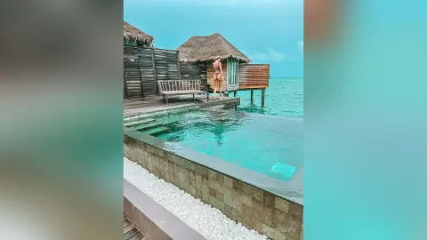 ChristineDavis stands naked on a wooden deck next to a clear blue swimming pool, overlooking the ocean near a thatched hut.
