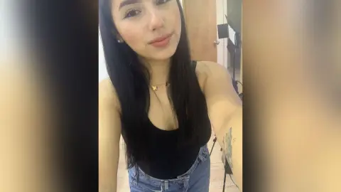 Smiling young woman with long dark hair wearing a black tank top and blue jeans in a bright indoor setting.