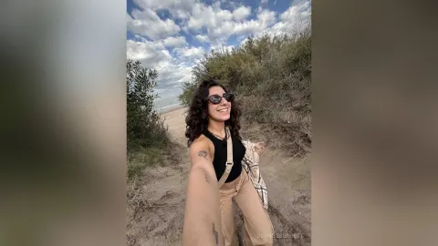 SophieeSunsett — SophieeSunsett smiles in sunglasses and a black tank top while standing on a sandy path near the ocean.
