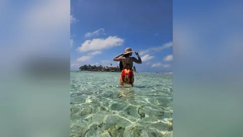 PamelaCreed stands in clear shallow ocean water wearing a red and yellow bikini and a sun hat, facing away from the camera toward a distant tropical island.