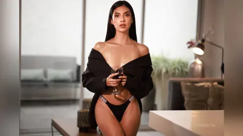 LucyMartin wears a black open shirt and black thong, posing while holding a glass of wine in a modern living room.
