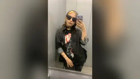 Edgy woman in black graphic shirt and sunglasses taking a moody mirror selfie in an elevator.