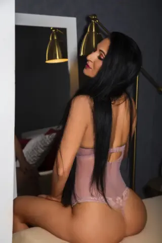 Woman with long black hair in a pink lace bodysuit sitting in a moody, dimly lit bedroom setting.