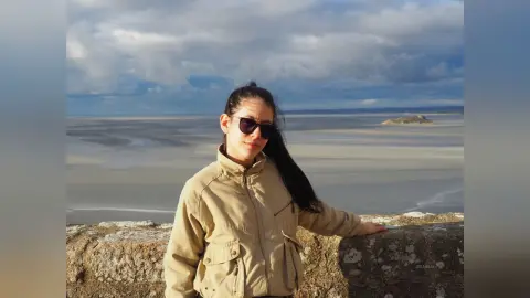 JessieLiu wears dark sunglasses and a tan jacket while standing outdoors against a coastal landscape or beach background.