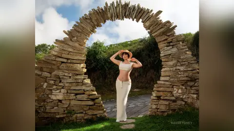 Fashionable woman in a white crop top and maxi skirt posing under a rustic stone archway in a sunny garden.