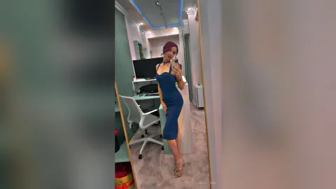 Woman in a blue sleeveless midi dress taking a mirror selfie in a modern bright room.