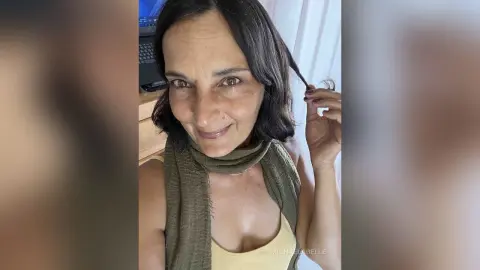 MichaelaBelle smiles while touching a strand of her dark hair, wearing a light yellow tank top and an olive green scarf.