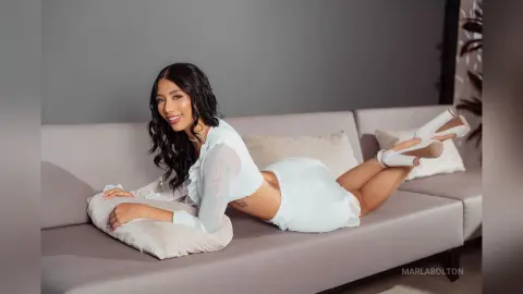 Smiling woman in a white summer outfit relaxing on a grey sofa in a bright, minimalist living room.