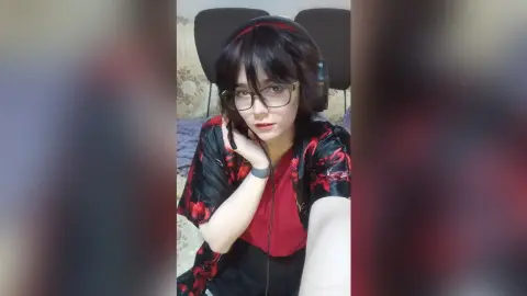 Young woman with glasses and headphones wearing a red and black floral kimono in a cozy bedroom setting.