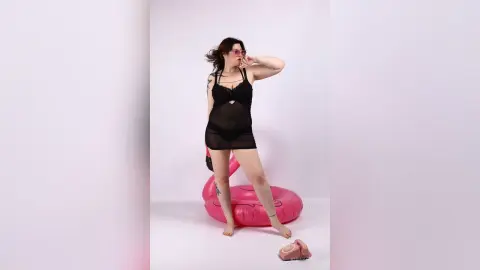KiraKovall wears a black sheer teddy and pink sunglasses while standing next a pink inflatable ring in a white studio.