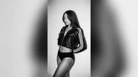 Stylish woman in a black leather Adidas jacket and briefs posing against a white wall in moody black and white.