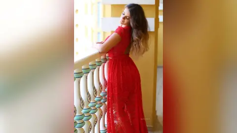 Elegant woman in a long red lace gown posing on a sunny balcony with a dreamy mood.
