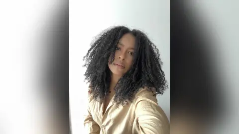 Smiling woman with voluminous curly hair wearing a beige linen shirt against a white background.