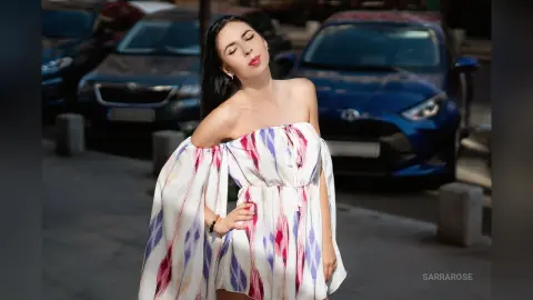 Stylish woman in a white off-the-shoulder floral sundress posing outdoors on a sunny street near parked cars.