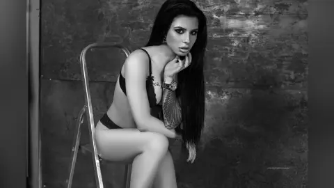 Moody black and white portrait of a long-haired woman in black lingerie sitting on a chair in a studio.