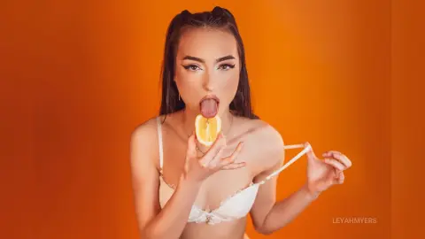 Young woman in white lace lingerie licking an orange slice against a vibrant orange background.