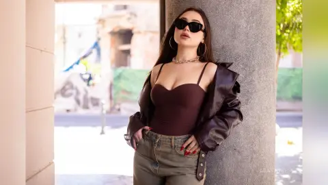 Stylish woman in a brown bodysuit, leather jacket, and sunglasses leaning against a pillar in a sunny urban setting.