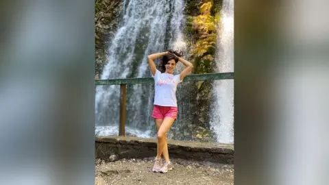 Cheerful woman in a white t-shirt and pink shorts posing in front of a scenic forest waterfall.