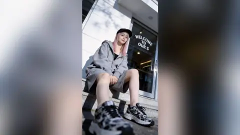 Young woman with pink hair in a grey hoodie and black sneakers sitting on urban concrete steps.
