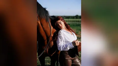 Redheaded woman in a white blouse and brown leather pants leaning against a brown horse in a sunny field.
