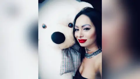 DeeaAnderson poses closely next to a large white teddy bear, wearing red lipstick and a silver choker necklace.