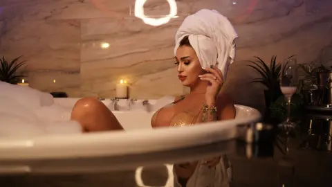 Relaxed woman in a white towel relaxing in a bubbly bathtub by candlelight in a luxury marble bathroom.
