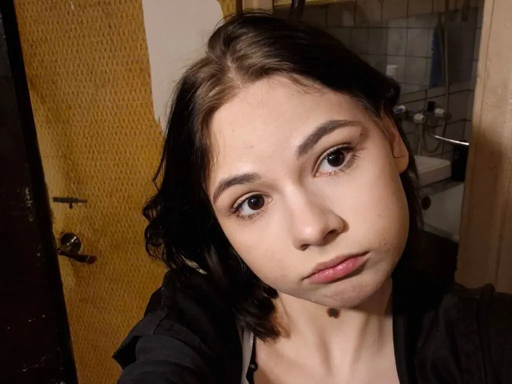 CeolaFilbert looks into the camera with a pouty expression, showing her dark hair and black shirt in a bathroom setting.