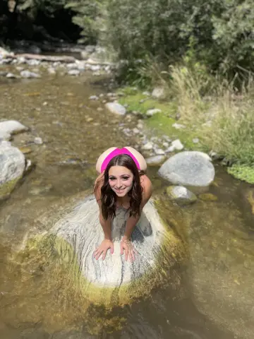 Princess1234, wearing a pink bikini, poses while leaning over a large rock in a shallow, clear stream surrounded by trees and greenery.
