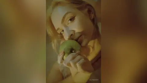 Blonde woman in a soft cream top biting a green apple in warm, moody indoor lighting.